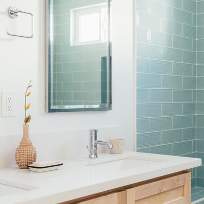 Bathroom with blue subway tile shower, vanity with white counter-wood cabinets, rectangular mirror and a vase with a dry plant cutting