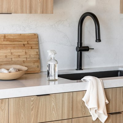 Spray bottle of cleaning liquid on kitchen counter with beige accent bowl and wood cutting board and black faucet sink