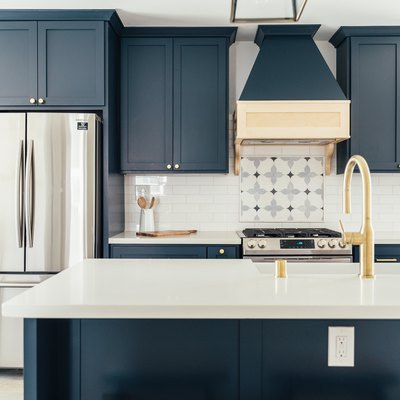 Dark blue kitchen cabinets with gold knobs. A silver French refrigerator. Ornate tile by stovetop. Kitchen island with gold sink faucet.