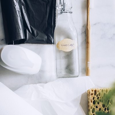 Black garbage bag, soap, empty glass bottle, wood toothbrush, paper towels, and sponger on white countertop