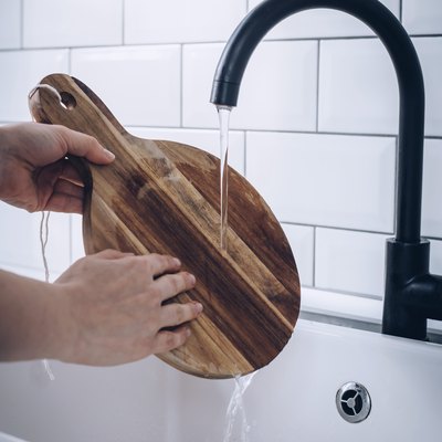 Hands washing round wood cutting board in white sink with black faucet against white tiled backsplash
