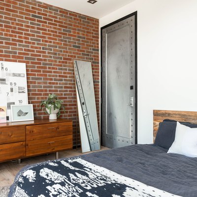 Wood cabinets with lamp, plant, and white diagram board against brick wall next to full-length mirror and black-and-white bed