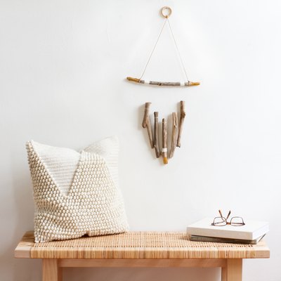 a wall hanging, made of several vertical pieces of driftwood suspended from a horizontal one, hangs above a wooden bench with a rattan top