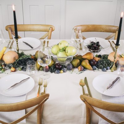 a line of produce, including grapes, pears, cherries, and squash runs down the length of a table with a white cloth and gold cutlery