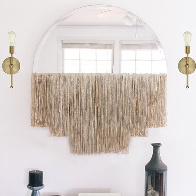 Round mirror with fringe beside gold sconces over white dresser with vase