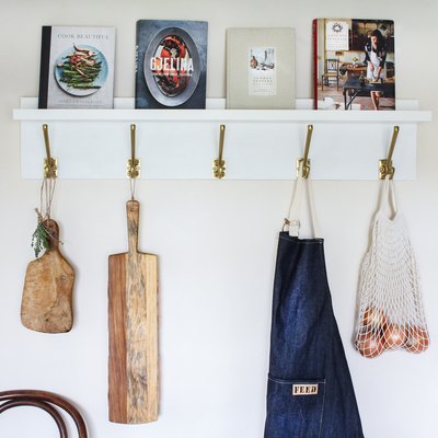 White kitchen wall hangers with gold hooks holding cutting boards, blue apron, and bag of tomatoes on white wall with books resting on top