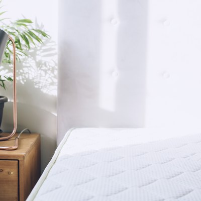 White mattress beside side table with small plant and lamp against white curtains.