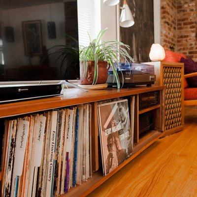A wood credenza with a record collection, tv and a plant, in a living room with a brick wall and wood floor.