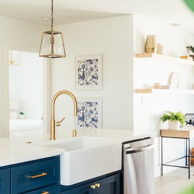 Dark blue kitchen island cabinets with gold hardware, white countertop, and gold sink faucet. Gold lantern pendant lights and wood shelves.
