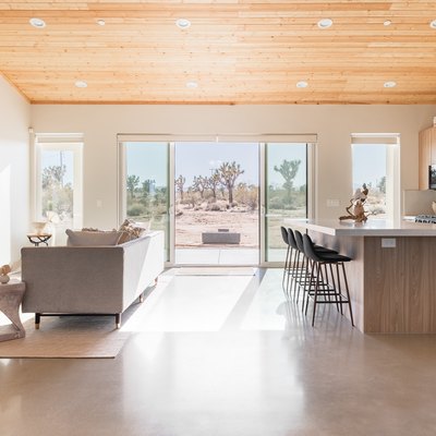 Minimalist wood cabinet kitchen and living room with gray furniture, sloped wood ceiling, and picture windows of desert
