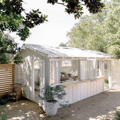 White greenhouse with a corrugated roof and open windows with wood counters. Wood fence and trees surround.