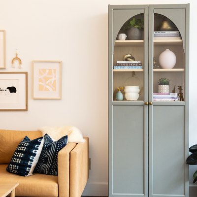 Gray cabinet with an arched top, inside are white vases, plants, and books and a beige sofa with blue pillows.