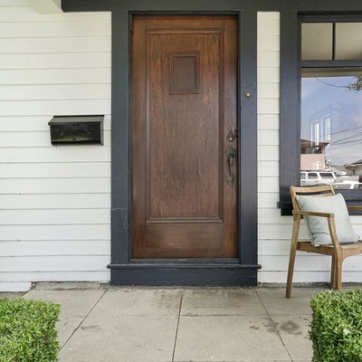 A wooden front door on a white house with a chair on the front porch