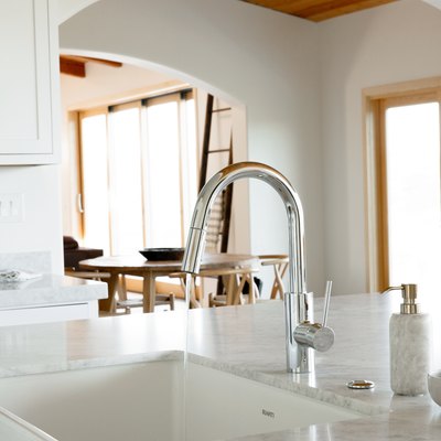 In a kitchen, a sink in a marble countertop, part of a kitchen island. The chrome faucet is running water. Next to the sink, a marble soap dispenser and a wooden scrub brush in a ceramic dish.