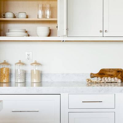 Kitchen counter. The countertop itself is marble, and the cabinets above and below are white. The top left cabinet is open, revealing a stacks of white plates,  white bowls, two white mugs, and two clear glasses. On the counter, three glass food storage containers, a cutting board, and a string of garlic.
