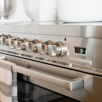 Closeup of an oven door and temperature control knobs on a stainless steel range. A white and grey striped kitchen towel is hanging over the handle over the oven door.