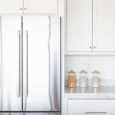 Double doors of a stainless steel refrigerator with white cabinets overhead. Next to it, marble countertop with glass food storage containers and white cabinets.