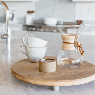 On a marble countertop, two stacked ceramic mugs, a Chemex, and a candle in a ceramic holder on a small round wooden board. In the background, a chrome faucet and a marbled soap dispenser.