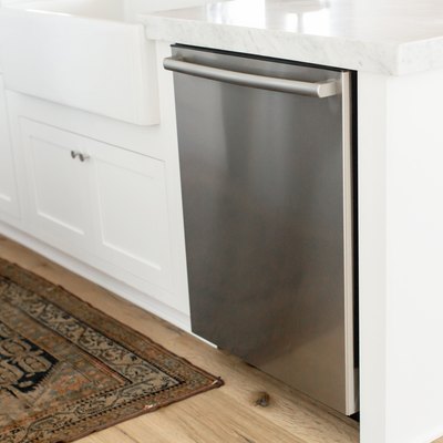 Stainless steel dishwasher in a white kitchen island. The marble countertop extends over the dishwasher. On the floor, a small rug.
