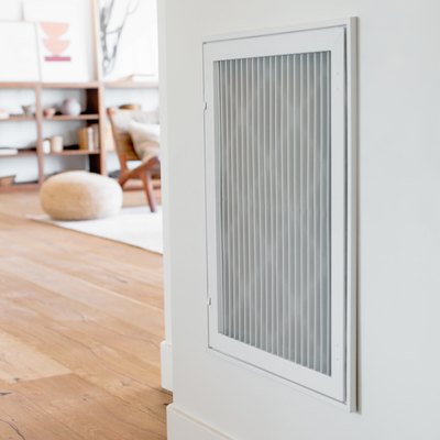 A white air vent on a white wall. Living room partially visible in the distance.