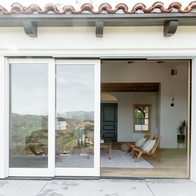 Glass sliding doors with white frame leading to a living room where two teak chairs with accent pillows and a rustic coffee table are visible.