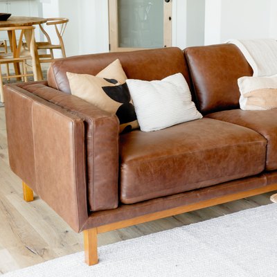 A brown leather sofa with three pillows and a white throw blanket. The sofa is on a white rug, and a wood coffee table is visible. Behind the sofa, there is a light wood table with four chairs and a dark brown bowl in the center of the table.