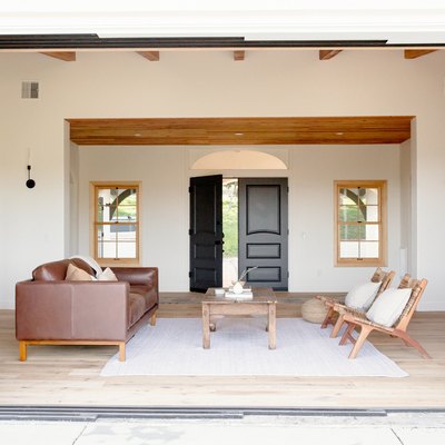 A large rectangular open doorway leading to a sitting room with a brown leather couch, a natural wood coffee table, and two wooden chairs with white pillows on a white rug. The walls of the room are white. Two windows and a dark wood double door are on the opposing wall.