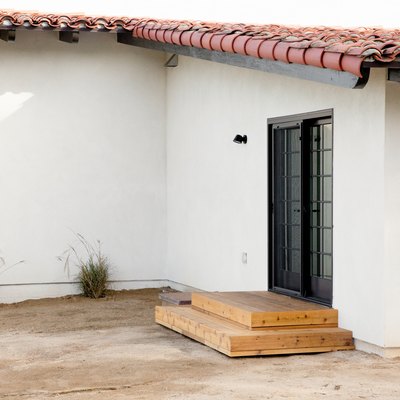 Back entrance to a white Spanish-style home with a clay tile roof. Small Mexican feather plants run along the side of the home. Two wooden steps lead up to a black sliding door.