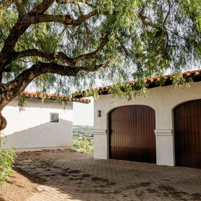 Brick driveway leading to a white Spanish-style garage with two wood doors. Oak tree by a driveway.