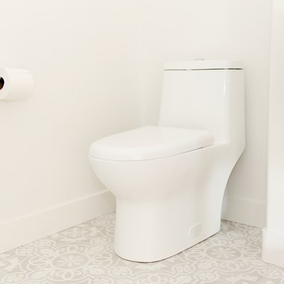 A white toilet in a bathroom with white walls and a white and grey tiled floor. A roll of toilet paper is on the wall next to the toilet, hanging on a simple chrome holder.