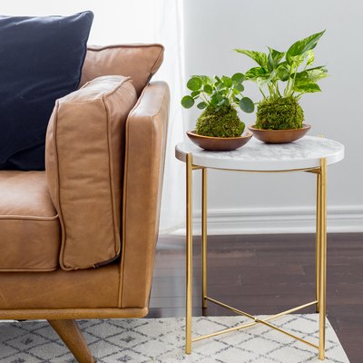 Marble side table with gold metal legs and two small potted plants beside tan leather couch in white-walled room with white rug.