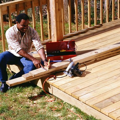 A carpenter sits on a deck measuring wood to complete a construction project.