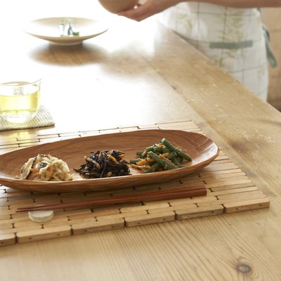 Japanese style vegetables in wooden tray with chopsticks on wooden table, woman preparing food in background