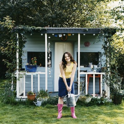 Woman Stood in Front of a Small Wooden Bungalow, Holding a Watering Can