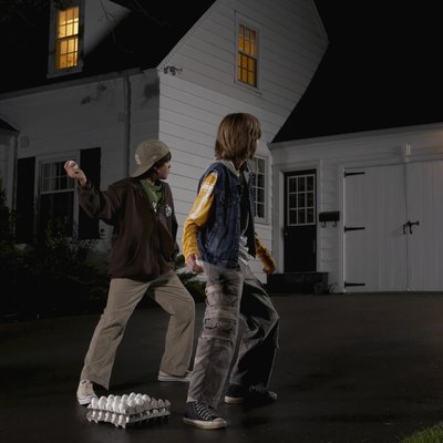 Two boys (12-13) standing in driveway, throwing eggs at house, night, rear view