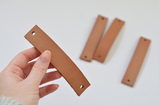 Hand holding piece of tan leather with a hole punched at each end.