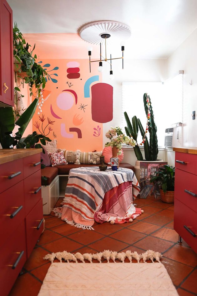 vermillion color kitchen cabinets, terracotta tile floor, art painted on wall, corner built in dining space, textiles over dining table, modern chandelier, plants.