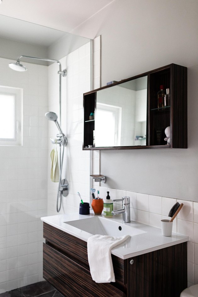 glass shower with white square tile, small wood vanity with white ceramic top, built-in medicine cabinet with mirror and wood shelves
