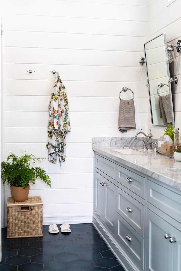 light blue painted bathroom cabinets, marble countertop, silver rectangular mirror, blue hexagon tile floor, rattan box, green plant, floral towel hung up