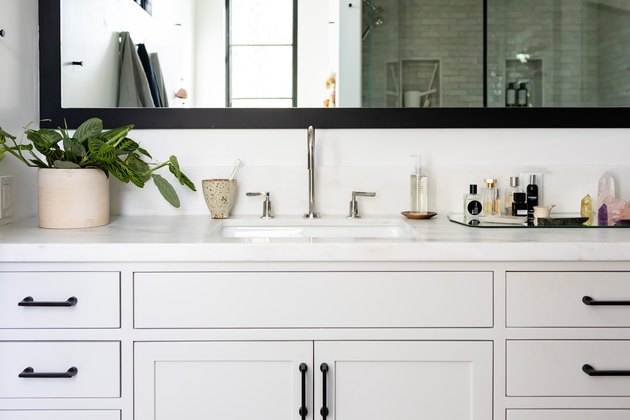 gray bathroom vanity cabinet, undermount sink, silver faucet and handles, rectangular mirror with black trim, vase with green plant, mirrored tray with various bathroom products