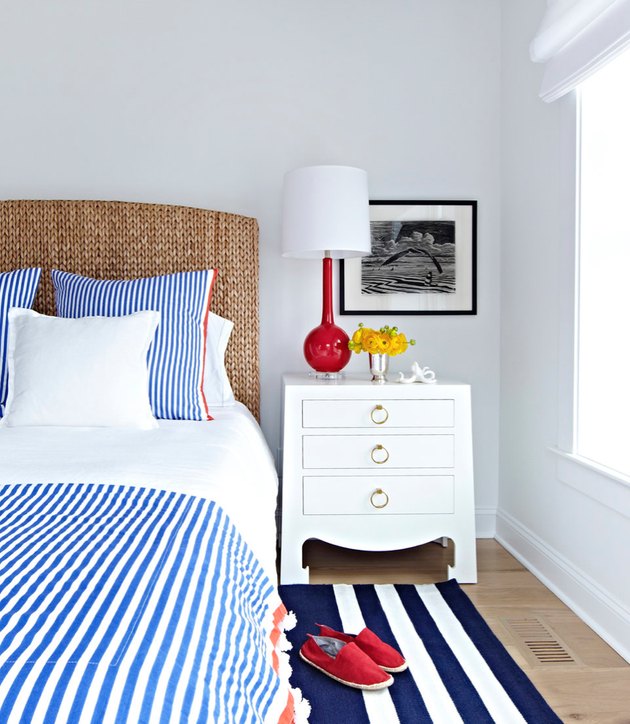 Blue and white striped shams and duvet, wicker headboard, and vermillion color table lamp with white shade, white night stand, black and white photograph, dark blue and white striped area rug, red shoes.