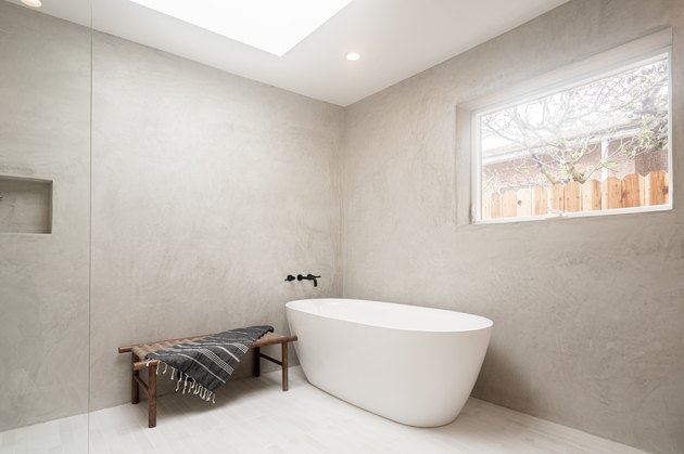 white freestanding tub in open shower space, wall-mounted black faucet and handles, small brown bench with towel draped over, glass shower door