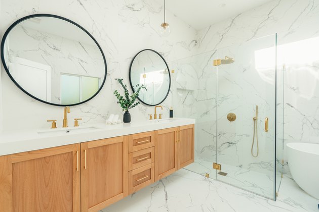 Bathroom with marble floor and walls, wood sink area, glass shower, large round mirrors