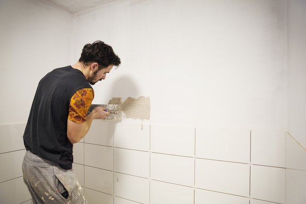A builder, tiler placing white ceramic tiles on a wall in a bathroom.