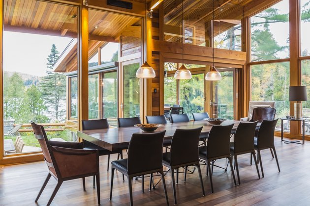 Wooden dining table with rawhide armchairs, black leather chairs and illuminated industrial style copper with frosted glass pendant lighting fixtures in dining area of great room with Ipe wood floor inside luxurious stained cedar and timber wood home wi