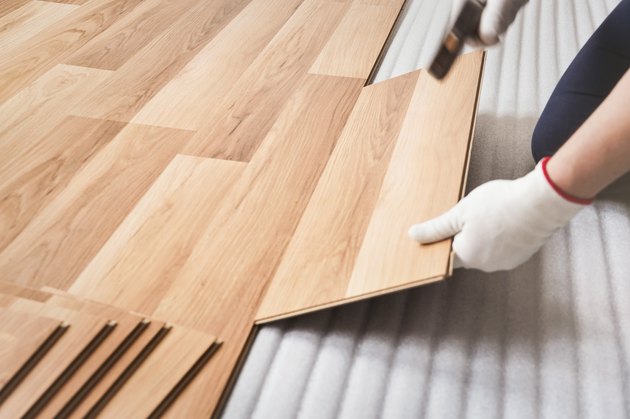 Installing laminated floor, man hand in white glove holding wooden tile and using hammer, over white foam base layer