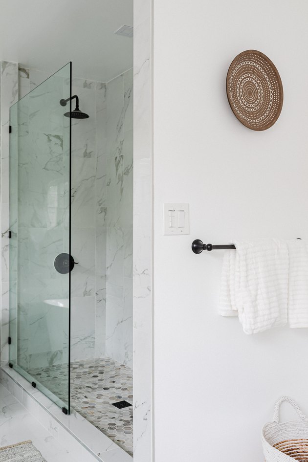 A glass shower in a white walled bathroom with a woven plate wall hanging