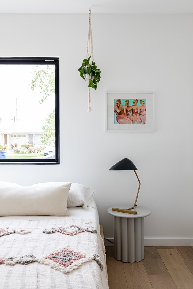 Bedroom with side table with lamp, white bed with pillow and quilted blanket, hardwood floor, white wall with window, hanging plant, and painting