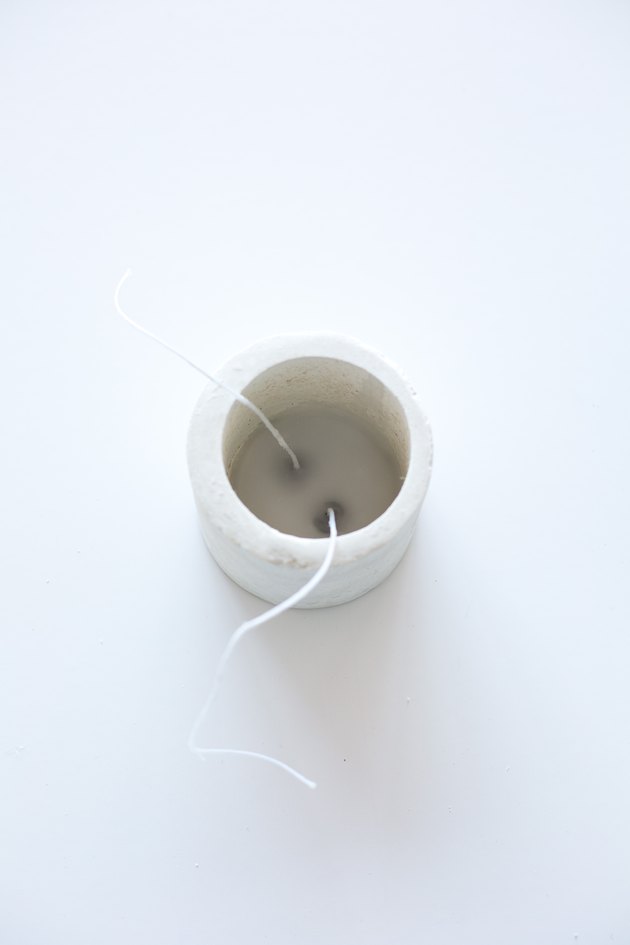 Two wicks partially submerged in wax in concrete candle holder on white background