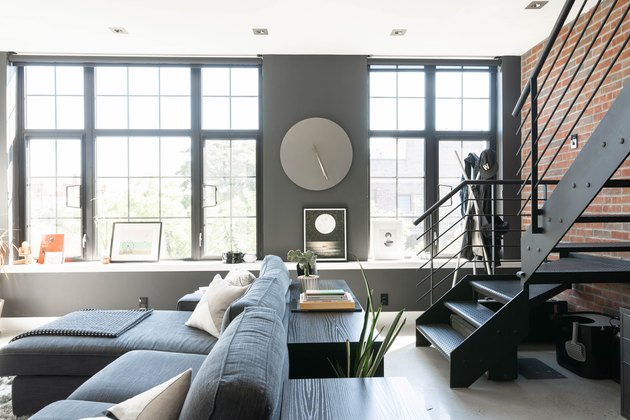 Modern upstairs living space with brick wall, grey couch with white pillows, full-length windows, couch, and long wood table